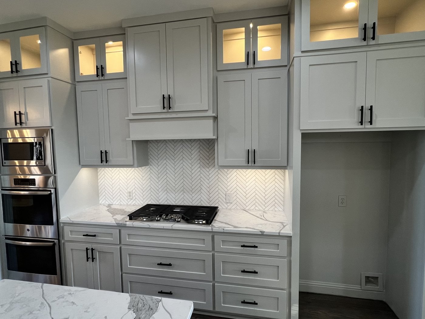 Gray kitchen cabinets with herringbone marble backsplash, gas cooktop and lit glass uppers
